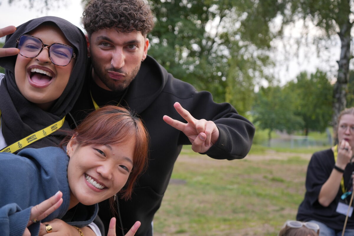 Drei Menschen schauen aus der linken unteren Ecke in die Kamera und grinsen